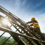 Worker in safety gear climbing a metal roof frame under sunlight.