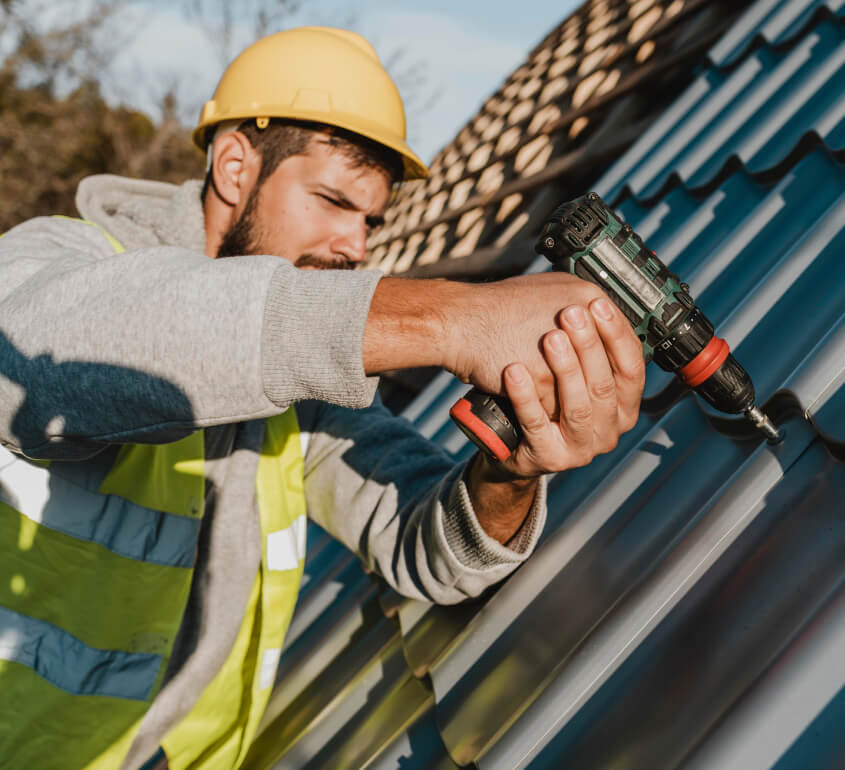 Construction worker in a yellow hard hat and safety vest using a power drill to install metal roofing panels on a sloped roof.