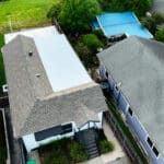 Aerial view of residential homes with different roofing types, including a flat white roof and pitched shingle roofs, surrounded by greenery and nearby structures.