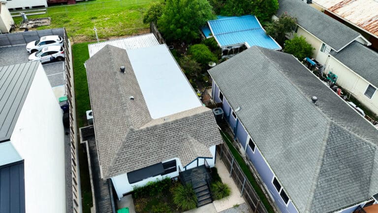Aerial view of residential homes with different roofing types, including a flat white roof and pitched shingle roofs, surrounded by greenery and nearby structures.