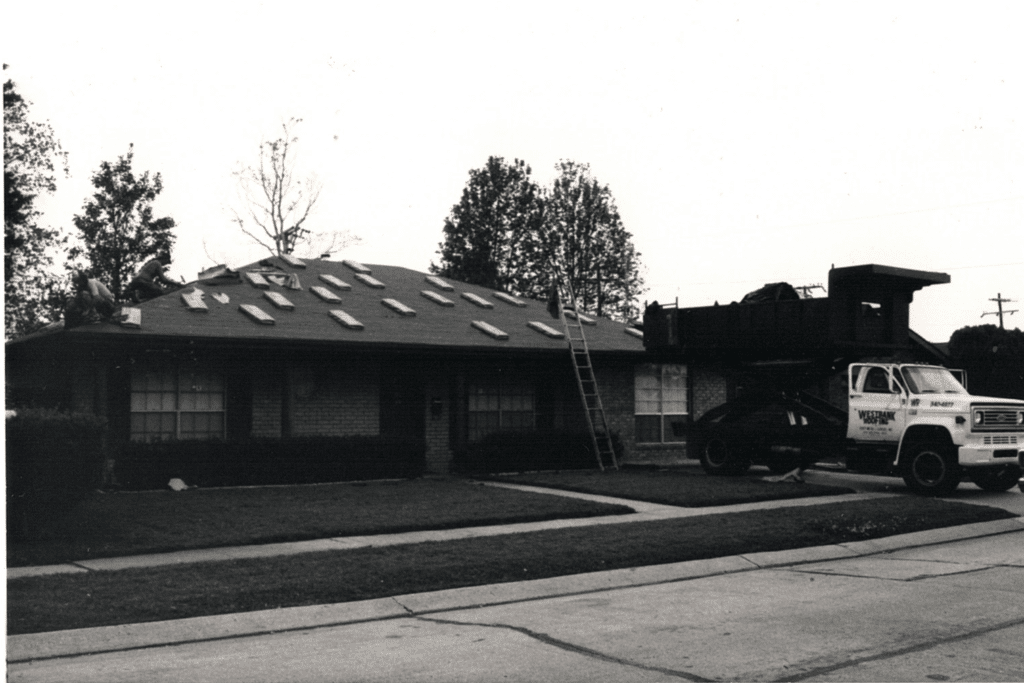 Black and white photo of a residential roofing job in progress, showing a house with shingles removed, a ladder leaning against the roof, and a roofing truck parked nearby.