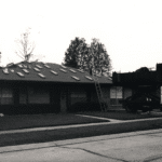 Black and white photo of a residential roofing job in progress, showing a house with shingles removed, a ladder leaning against the roof, and a roofing truck parked nearby.