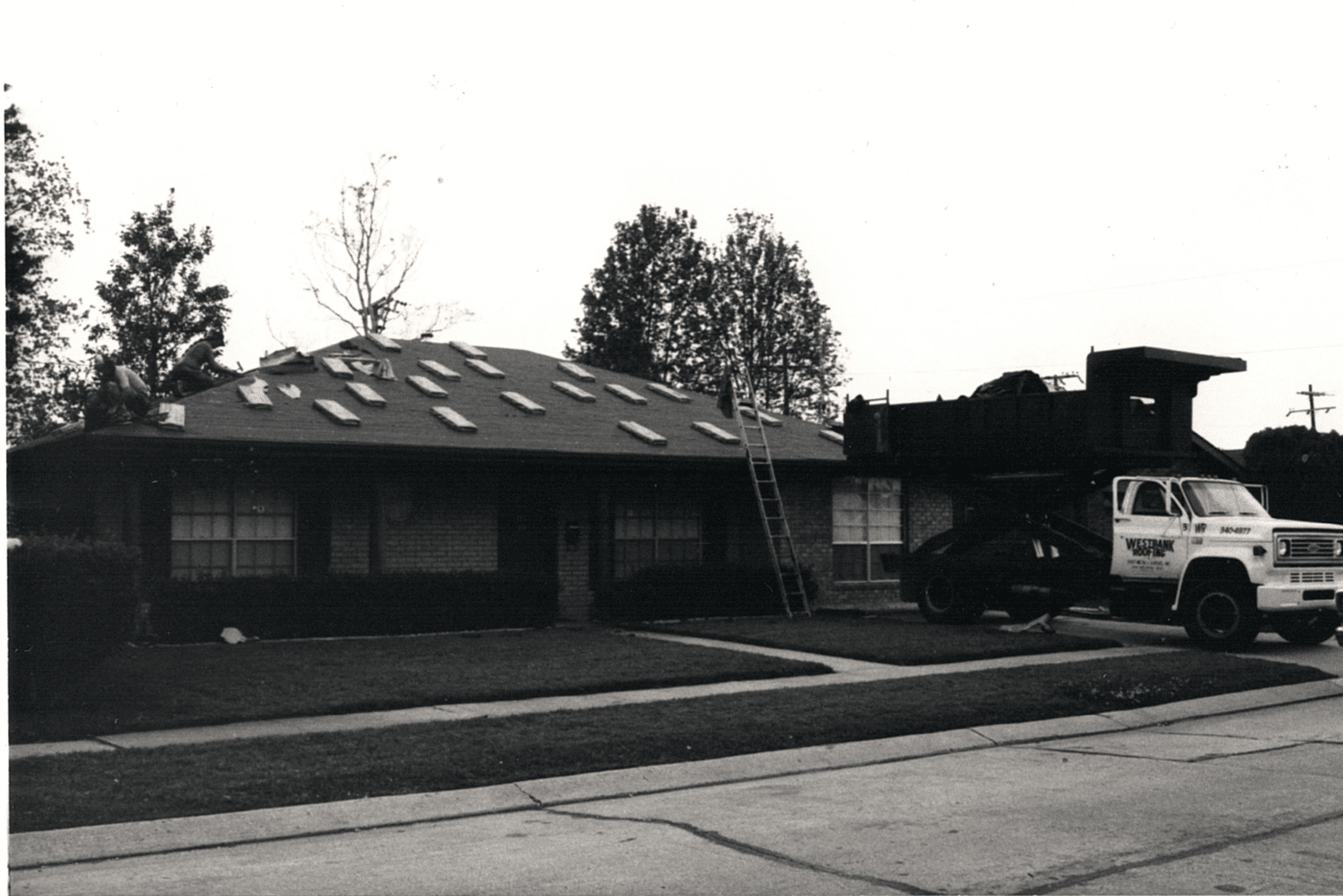 Black and white photo of a residential roofing job in progress, showing a house with shingles removed, a ladder leaning against the roof, and a roofing truck parked nearby.
