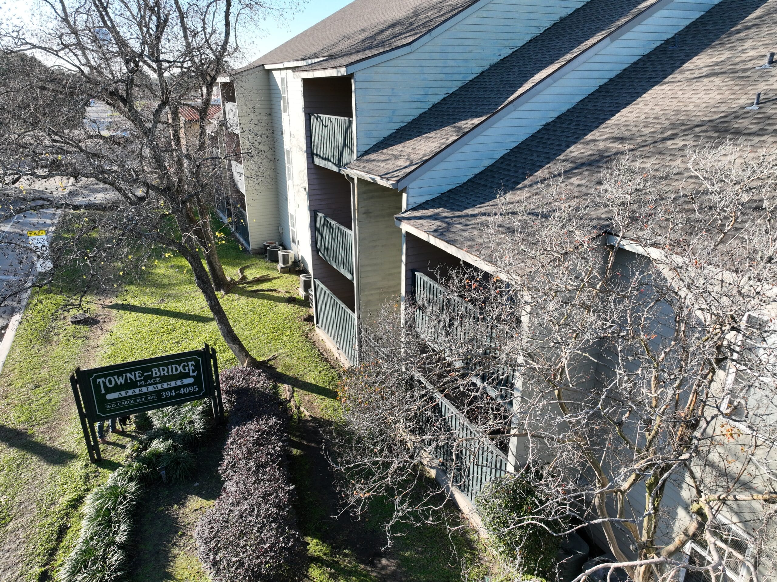 Aerial view of the Towne-Bridge Place Apartments sign and building exterior, featuring balconies, bare trees, and a neatly landscaped walkway.